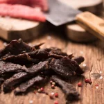 Beef jerky and spices on a wood table with raw beef on a cutting board.